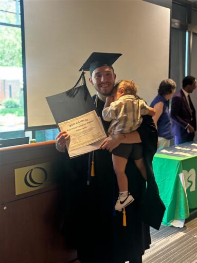 Josh in a cap and gown, holding a baby on one shoulder, proudly showing his diploma