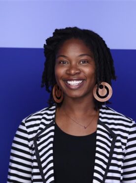 Kia wearing a striped blazer, copper colored circular earrings, smiling at the camera